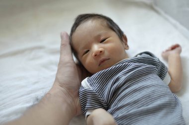 Bir bebek kolları açık beyaz bir çarşafın üzerinde yatıyor. Bebeğin üzerinde siyah beyaz çizgili bir gömlek var.