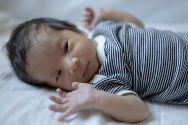 Bir bebek kolları açık beyaz bir çarşafın üzerinde yatıyor. Bebeğin üzerinde siyah beyaz çizgili bir gömlek var.