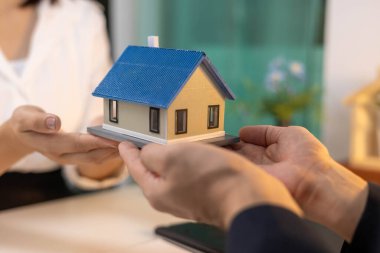 Bir adam elinde model bir ev tutuyor. Arkasında bir kadın var. Kadın beyaz gömlek ve siyah pantolon giyiyor.