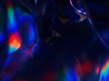 Renkli ışık yansımalarının soyut yakın çekimi. Görüntü, derinlik ve hareket hissi uyandıran canlı ve yanardöner renklere sahip koyu bir arkaplan içeriyor..