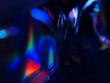 Renkli ışık yansımalarının soyut yakın çekimi. Görüntü, derinlik ve hareket hissi uyandıran canlı ve yanardöner renklere sahip koyu bir arkaplan içeriyor..