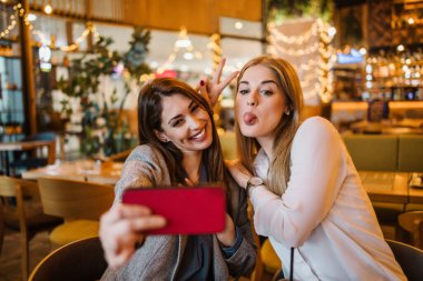 Alışveriş yaptıktan sonra kafeteryada ya da restoranda oturup mutlu mesut sohbet eden iki arkadaş. Selfie çekmek için akıllı telefon kullanıyorlar..