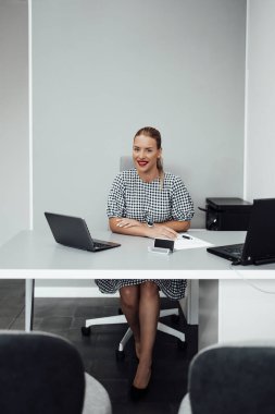 Young confident woman sitting in her office and looking at camera. Plain gray wall in background.