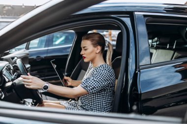 Beautiful young woman working as used car seller. She is using digital tablet and checking car condition before customer or buyer. Used vehicle dealership.