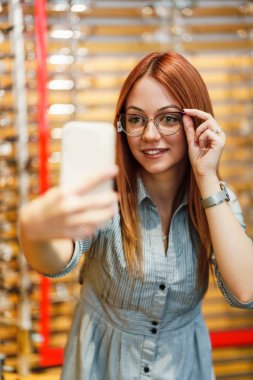 Beautiful and fashionable redhead woman choosing eyeglasses frame in a modern optical store. She is happy and satisfied with her glasses and takes a selfie photo with smart phone.