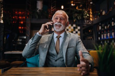 Restoranda oturup öğle yemeğini bekleyen mutlu iş adamı. Akıllı telefon kullanıyor ve biriyle konuşuyor. İş dünyası yaşlıları yaşam tarzı konsepti.