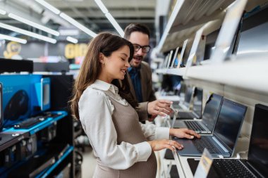 Beautiful and happy middle age couple buying consumer tech products in modern home electronics store. They are choosing and trying laptop computer. People and consumerism concept.