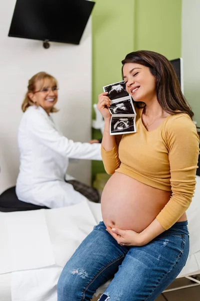 Beautiful and pregnant woman is happy after pregnancy medical check up. She is holding and looking at ultrasound scan of her baby. Modern pregnancy healthcare concept.