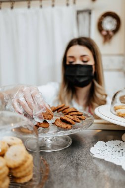 Koruyucu maskeli güzel bir bayan satıcı bir şekerleme dükkanında ya da fırında çalışıyor. Lezzetli tatlılar satıyor ve paketliyor. Coronavirus ve küçük işletme konsepti.