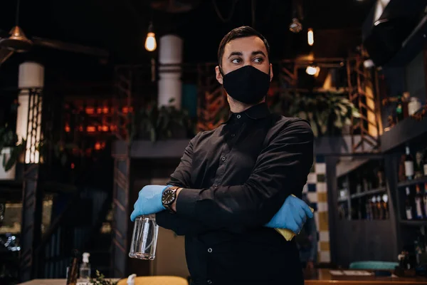 Portrait of a serious waiter standing in a nice restaurant. He wears a protective mask and gloves as part of security measures against the Coronavirus pandemic.