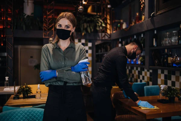Young restaurant waiters cleaning and disinfecting tables and surfaces during Coronavirus pandemic disease. They are wearing protective face masks and gloves.