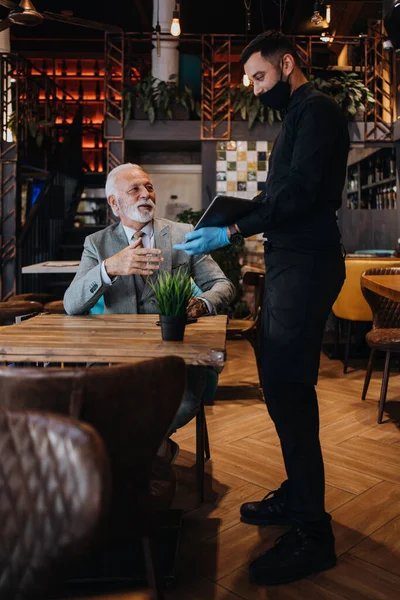 The waiter taking order for a delicious meal to the senior businessman at the restaurant. He wears a protective mask as part of security measures against the Coronavirus pandemic.