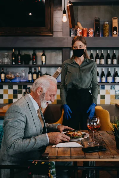 Waitress serves and takes the order from the senior businessman at the restaurant. She wears a protective mask as part of security measures against the Coronavirus pandemic.