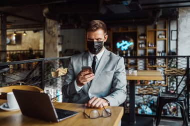 Yüzünde koruyucu maske olan bir iş adamı bir kafede oturuyor ve dizüstü bilgisayar kullanırken telefonla konuşuyor. Coronavirus, Covid-19 konsepti.
