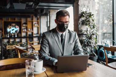 Yüzünde koruyucu maske olan bir iş adamı bir kafede oturuyor ve dizüstü bilgisayar kullanırken telefonla konuşuyor. Coronavirus, Covid-19 konsepti.