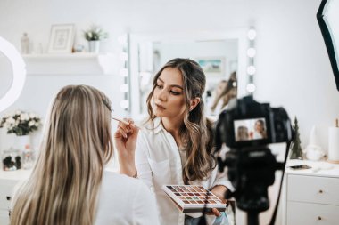 Evden ya da atölyeden güzel makyaj videosu yayınlayan mutlu genç bir kadın. Güzel çevrimiçi içerik kozmetikçisi makyaj yapıyor ve bazı iş araçlarını açıklıyor. Günlük kaydı ve çevrimiçi kanal çalışması. 