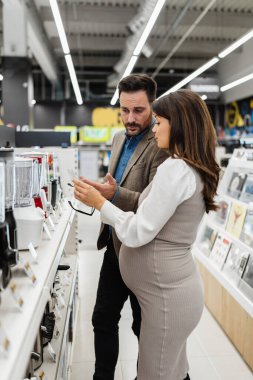 Beautiful and happy middle age couple buying consumer tech products in modern home tech store. They are choosing small kitchen appliances. People and consumerism concept.