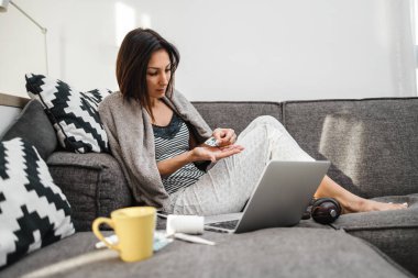 Hasta ve yorgun orta yaşlı kadın. Grip ve baş ağrısı çeken serbest çalışan. Haplar alıyor, bej bir battaniyeye sarılı olarak evdeki kanepede oturuyor..