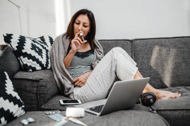 Hasta ve yorgun orta yaşlı kadın. Grip ve baş ağrısı çeken serbest çalışan. Haplar alıyor, bej bir battaniyeye sarılı olarak evdeki kanepede oturuyor..