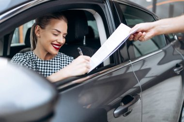 Satisfied and smiled female buyer sitting in her new car. She happy while signing buyer's purchase contract with used car seller.