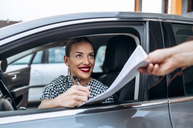 Yeni arabasında oturan tatmin olmuş ve gülümseyen bayan alıcı. İkinci el araba satıcısıyla alıcı sözleşmesi imzalarken mutlu oluyor..