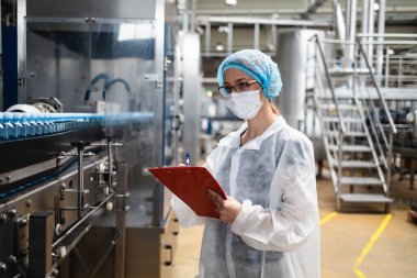 Koruyucu iş kıyafetlerinde çalışan, tıbbi malzeme araştırma ve üretim fabrikasında çalışan kadın işçi. Kontrol köprüsünde duruyor ve üretim hattını izliyor. Denetim kalite denetimi.