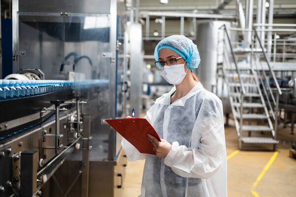 Женщина-работница в защитной рабочей одежде работает на научно-исследовательской и производственной фабрике медицинских товаров. Она стоит на мосту управления и наблюдает за производственной линией. Контроль качества.
