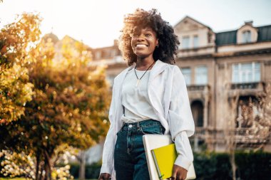 Güzel ve mutlu Afrikalı kız öğrenci üniversite kampüsünde dışarıda eğleniyor. O, dizüstü bilgisayarını tutarken ve üniversite parkında yürürken mutlu ve gülümsüyor. Arka plandaki eski bina.