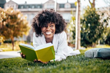 Güzel ve ciddi bir Afrikalı kız öğrenci fakülte sınavına çalışırken üniversite kampüsünde eğleniyor. Çimlerin üzerinde uzanıyor ve bir şeyler okuyor. Arka plandaki eski bina.