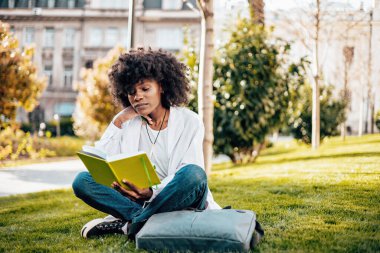 Güzel ve ciddi bir Afrikalı kız öğrenci fakülte sınavına çalışırken üniversite kampüsünde eğleniyor. Çimlerin üzerinde uzanıyor ve bir şeyler okuyor. Arka plandaki eski bina.