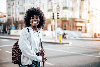Güzel Afrikalı genç kız öğrenci ya da turist şehir caddesinde duruyor ve toplu taşıma için bekliyor. O mutlu ve gülümsedi. Alanın kısa derinliği.