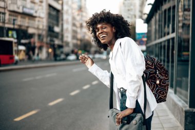 Güzel Afrikalı genç kız öğrenci ya da turist şehir caddesinde duruyor ve toplu taşıma için bekliyor. O mutlu ve gülümsedi. Alanın kısa derinliği.