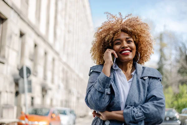 美丽的黑人妇女走在街上 用智能手机进行交流 她很高兴 阳光灿烂的日子 — 图库照片