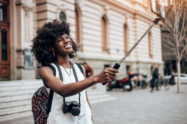 Genç siyah bayan turist güzel bir Avrupa şehrinin sokaklarında yürümekten zevk alıyor. Mutlu ve akıllı telefonunu selfie portreleri ve videolar çekmek için kullanıyor. Parlak güneşli bir gün.