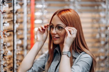 Beautiful and fashionable redhead woman choosing eyeglasses frame in a modern optical store.