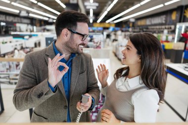 Beautiful and happy middle age couple buying consumer tech products in modern home appliances store. They are choosing beard and hair clipper and hairdryer. People and consumerism concept.