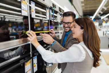 Beautiful and happy middle age couple buying consumer tech products in modern home appliances store. They are choosing electrical cooker and oven. People and consumerism concept.
