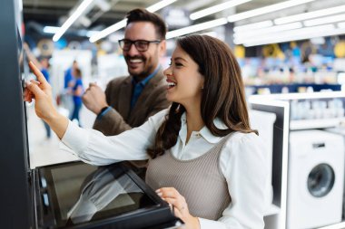 Beautiful and happy middle age couple buying consumer tech products in modern home appliances store. They are choosing electrical cooker and oven. People and consumerism concept.