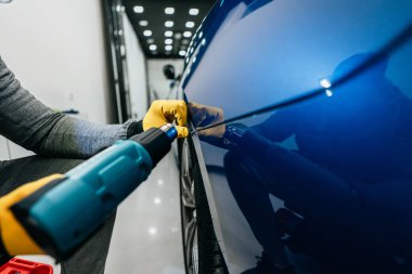 Modern bir araba atölyesinde profesyonel araç temizleme servisi. Araba atölyesi uzmanı arabaya vinil folyo ya da film koyuyor. İşçi folyo kenarlarını daha iyi sarmak ve sıkmak için buhar tabancası kullanıyor. 