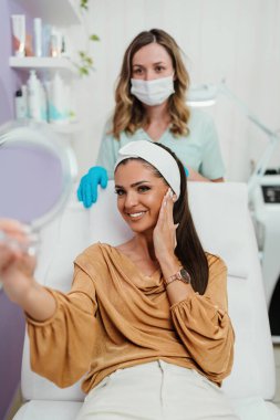 Güzel ve mutlu genç bir kadın tıbbi sandalyede oturup aynaya bakıyor. Hyaluronik asit doldurucu ya da botulinyum toksin iğneleriyle başarılı bir güzellik tedavisinden sonra tatmin oluyor..