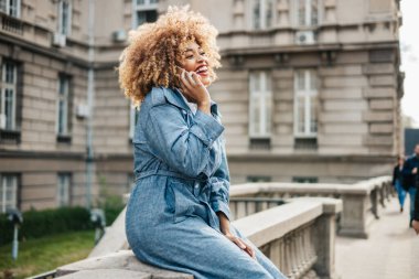 Güzel siyah kadın sokakta yürüyor ve iletişim için akıllı telefonu kullanıyor. O mutlu ve gülümsedi. Parlak güneşli bir gün.