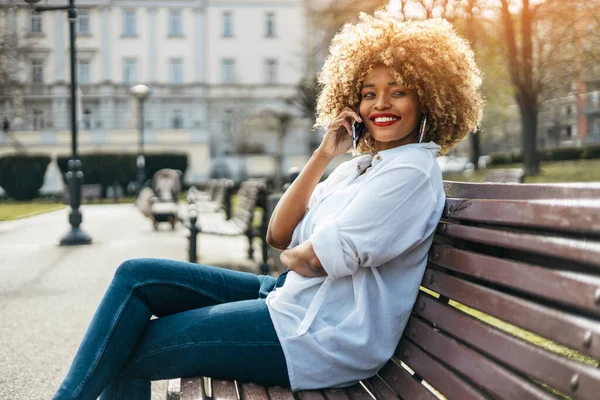 漂亮的黑人妇女坐在街头长椅上 用智能手机进行在线交流 她很高兴 阳光灿烂的日子 — 图库照片