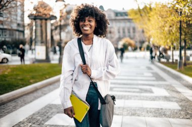 Güzel ve mutlu siyah kız öğrenci üniversite kampüsünde dışarıda eğleniyor. O, dizüstü bilgisayarını tutarken ve üniversite parkında yürürken mutlu ve gülümsüyor. Arka plandaki eski bina.