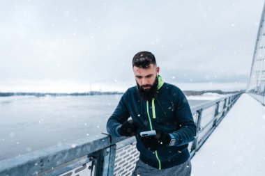 Çok soğuk ve karlı bir günde sakallı, koşan ve dışarıda egzersiz yapan yakışıklı orta yaşlı bir adam. Spor ve spor motivasyon teması. Aktivite verilerini takip etmek için akıllı telefon kullanıyor..