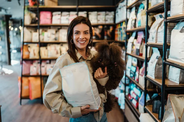 彼女の愛らしい茶色のおもちゃのプードルと一緒に現代ペットショップで楽しんでいる美しい若い女性 彼女は犬のフードバッグを保持しています — ストック写真