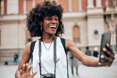Genç siyah bayan turist güzel bir Avrupa şehrinin sokaklarında yürümekten zevk alıyor. O mutlu ve akıllı telefonunu bazı fotoğraflar ve videolar çekmek için kullanıyor. Parlak güneşli bir gün.