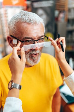 Tecrübeli kadın optometrist, yaşlı erkek hastasına göz bebeği mesafesini ölçüyor. Manüel gözbebeği kullanıyor..
