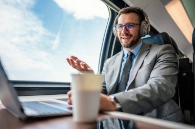 Yakışıklı iş adamı yüksek hızlı trenle seyahat ederken iyi vakit geçiriyor. Çevrimiçi iletişim, oyun ve eğlence için dizüstü bilgisayar ve kablosuz kulaklık kullanıyor.