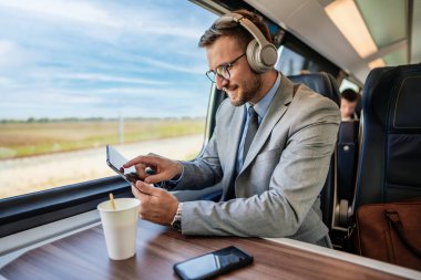Yakışıklı iş adamı yüksek hızlı trenle seyahat ederken iyi vakit geçiriyor. Çevrimiçi iletişim, oyun ve eğlence için dizüstü bilgisayar ve kablosuz kulaklık kullanıyor.
