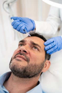 Yakışıklı orta yaşlı sakallı adam güzellik kliniğinde gençleştirici yüz enjeksiyonu yaptırıyor. Güzellik uzmanı erkek kırışıklıklarını botulinyum toksin iğneleriyle ya da hyaluronik asit doldurucularıyla dolduruyor..